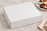 A white corrugated cardboard gift box shown on a light marble surface, with a plate of cupcakes and a striped napkin in the background.